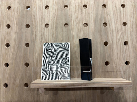 oak veneer Wooden pegboard with a shelf holding a black and white patterned box and a black rectangular peg against a wooden background.