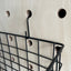 detail of black metal hook thread through hole of pegboard where the black metal wire basket is hanging off